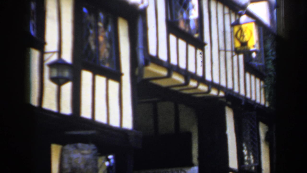 1949 England home-movie frame showing close timber-framed facades, white plaster panels and hanging lanterns along a narrow street.