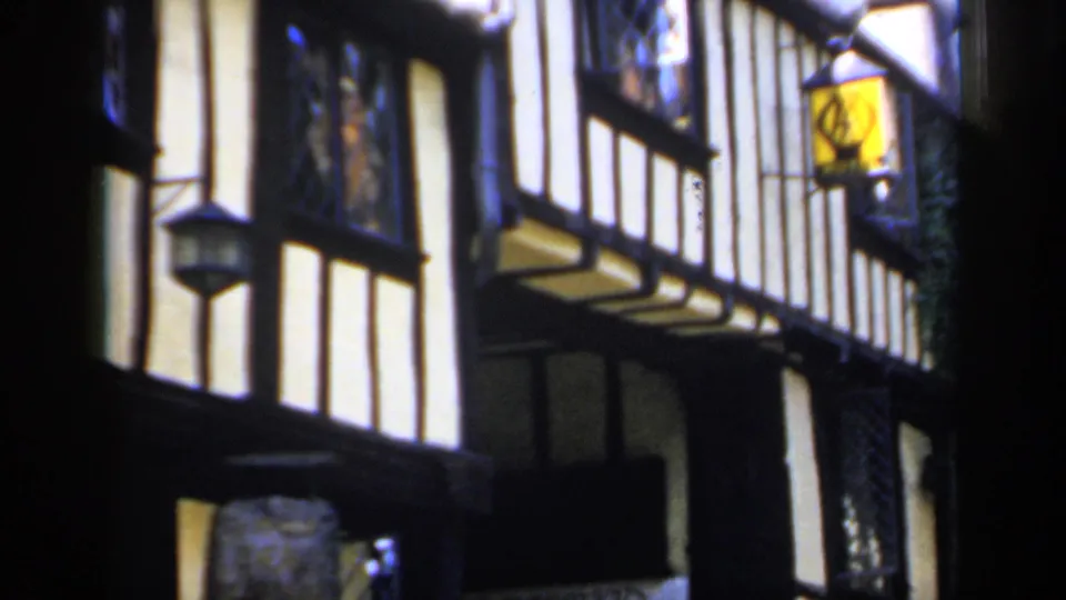 1949 England home-movie frame showing close timber-framed facades, white plaster panels and hanging lanterns along a narrow street.