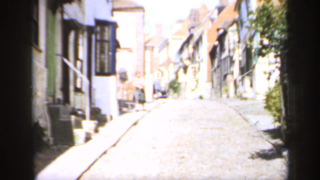 1949 England home-movie view down a bright cobblestone street with timber-framed buildings and older houses on both sides.
