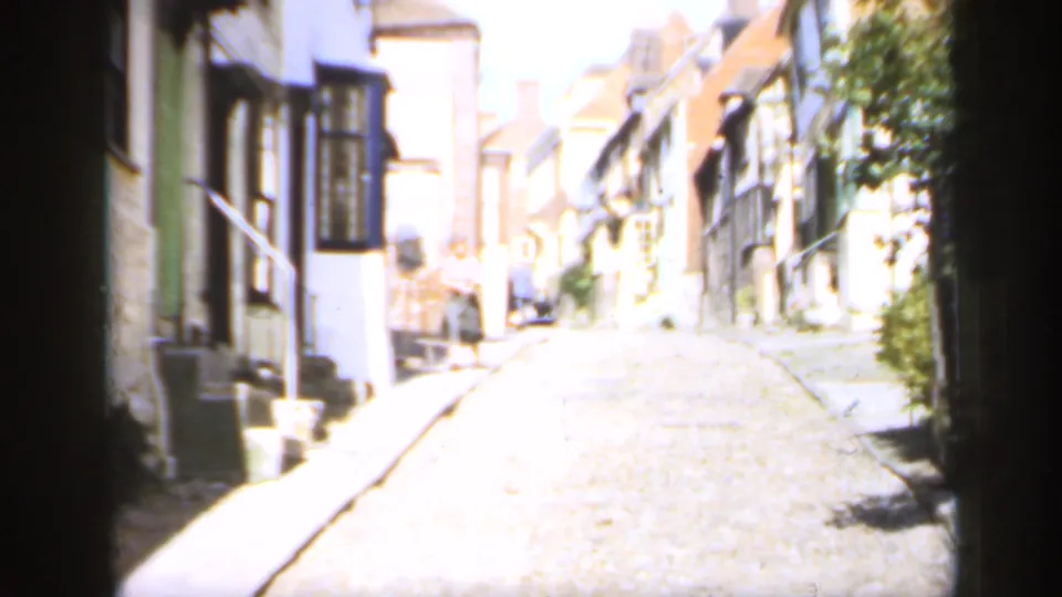 1949 England home-movie view down a bright cobblestone street with timber-framed buildings and older houses on both sides.