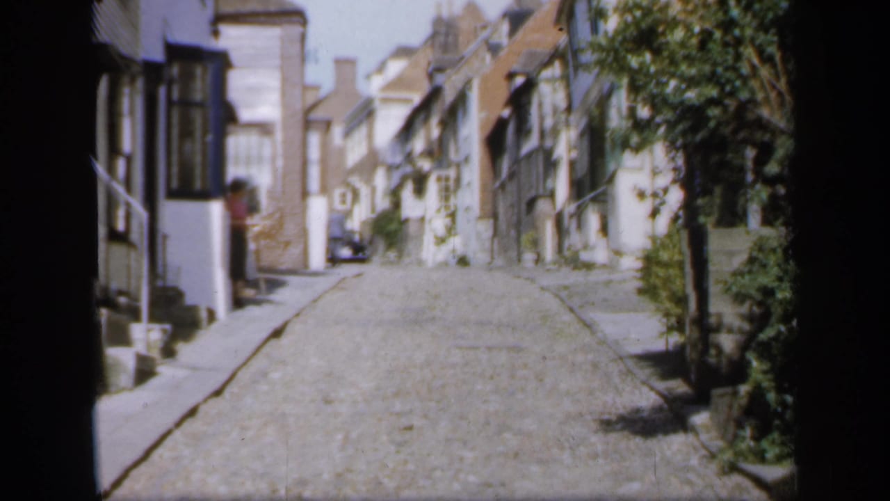 1949 England home-movie view of a narrow cobbled street with white and timbered houses, greenery, and a distant parked car.