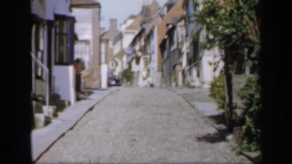 1949 England home-movie view of a narrow cobbled street with white and timbered houses, greenery, and a distant parked car.