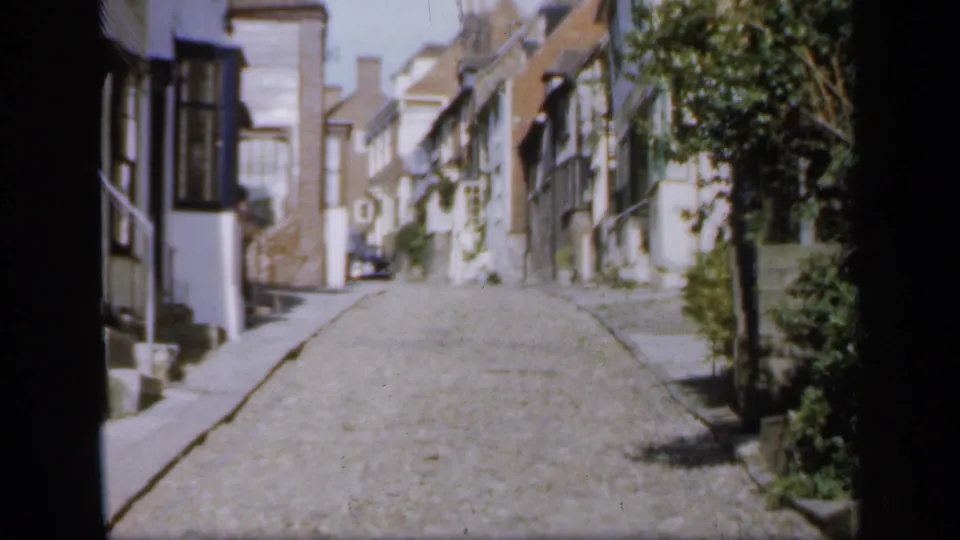 A 1949 English street scene shows cobbled paving, close-set houses, foliage, and a small car parked farther up the lane.