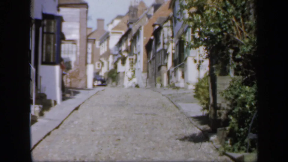 1949 England home-movie frame of a cobbled street where an adult appears partly visible near a doorway beside traditional houses.