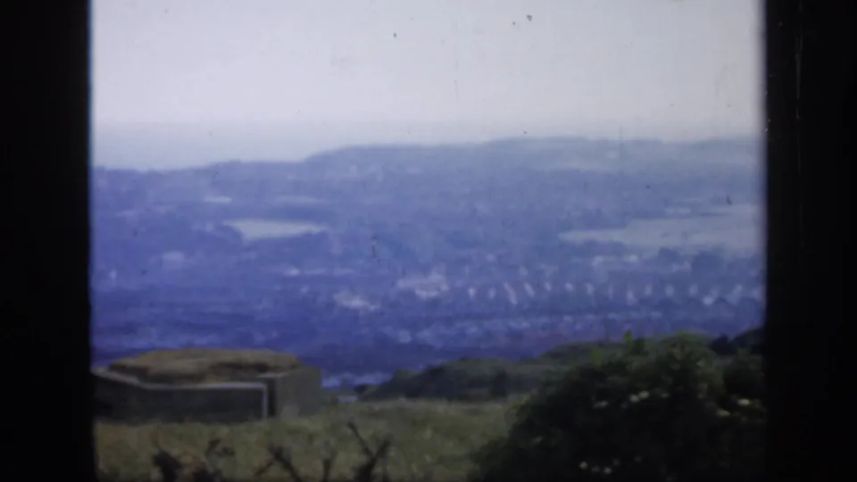 A high vantage view in England looks over a hazy town, fields, low hills, and possible coastline or broad water beyond.