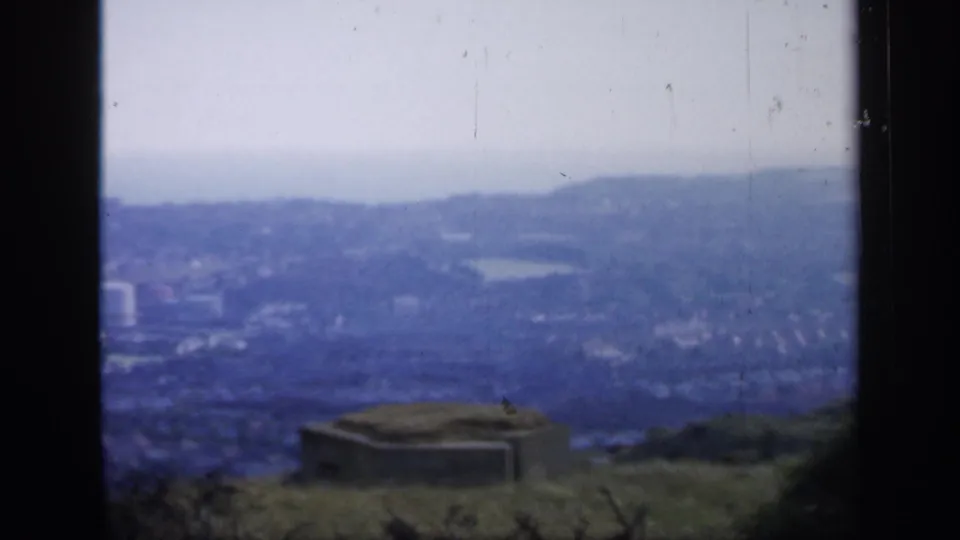 1949 England frame looking across a hazy town from above, with a low concrete structure on grass and dark window framing.