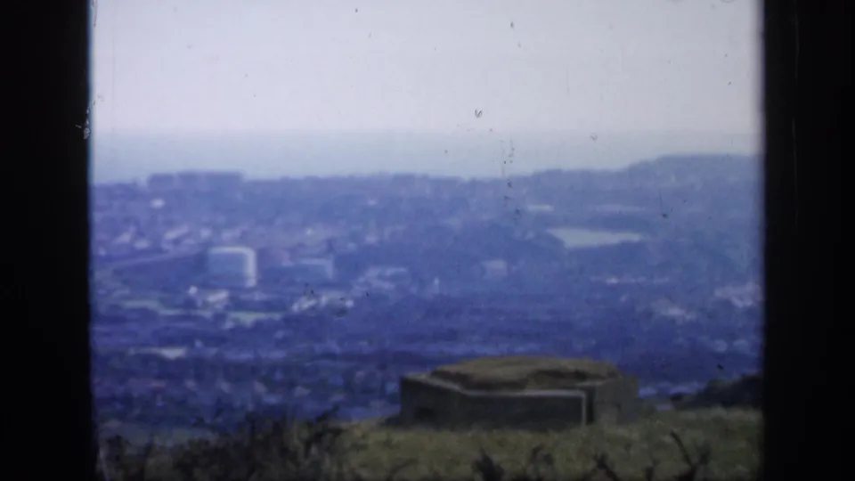 1949 England home-movie still with a hazy elevated town view, dark frame edges, grassy foreground, and low concrete structure.