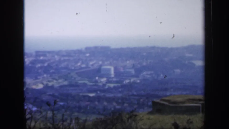 1949 England frame showing a hazy town panorama from a high vantage point, with foreground grass and a small concrete structure.