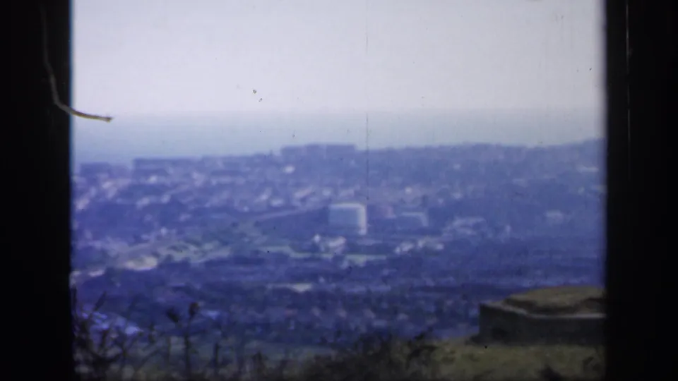 1949 England home-movie frame showing a hazy coastal town below a hill, bordered by dark window or gate edges.