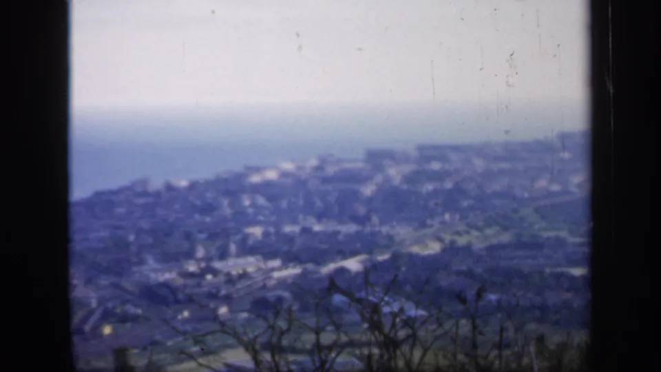 1949 England frame with a high view over houses and roads descending toward the sea, softened by haze and 8mm grain.