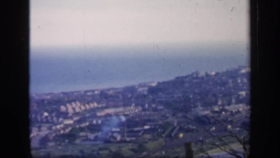 1949 England 8mm view across a coastal town, with rows of buildings, haze, and a faint smoke plume.