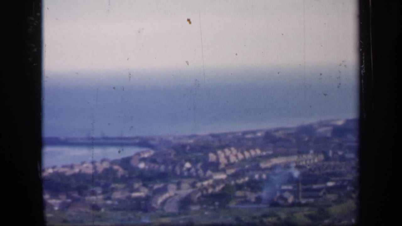 Soft coastal town view with sea, shoreline buildings, roads, and dark film-frame borders.