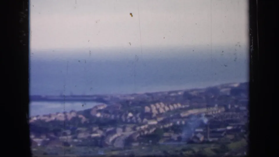 1949 England 8mm frame showing a hazy elevated view over a seaside town, shoreline roads, clustered buildings, and the sea beyond.