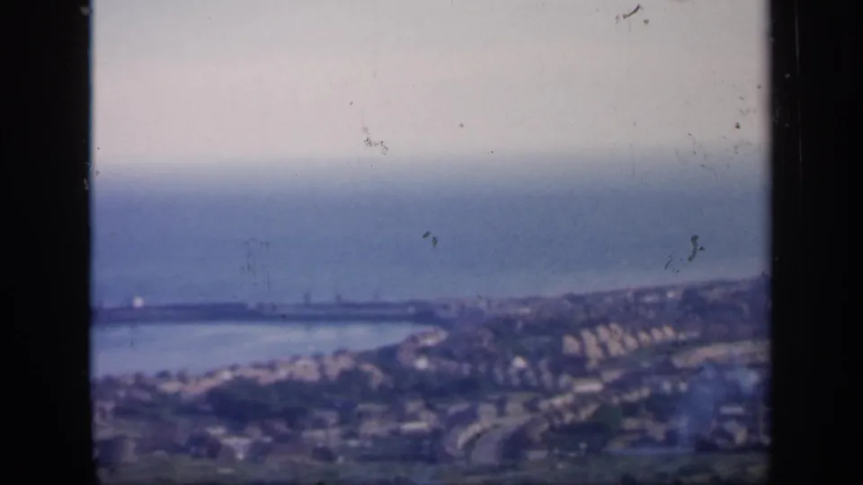 1949 England home-movie frame with a harbor area, sea horizon, shoreline houses, and soft focus from the 8mm scan.