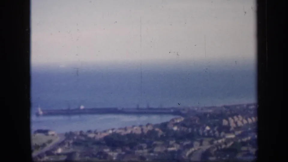 1949 England 8mm frame showing harbor water and a breakwater or pier, with a residential shoreline under a hazy sky.