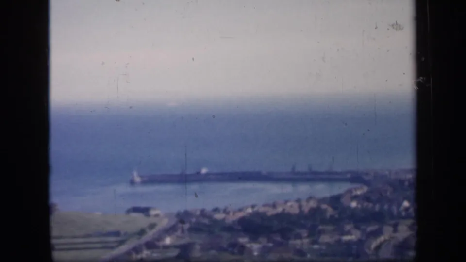 1949 England home-movie frame looking across green shoreline and harbor water toward the open sea and distant town edge.
