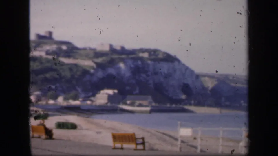 1949 England coastal frame with a promenade bench, railing, bay water, and chalk cliffs beyond.