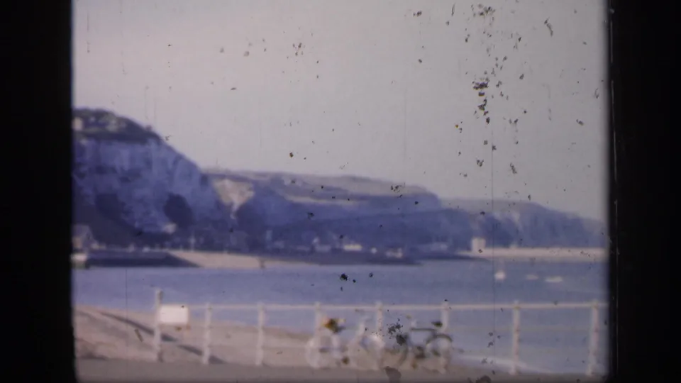 1949 England home-movie frame showing bicycles by a white seafront railing with calm water and chalk cliffs beyond.