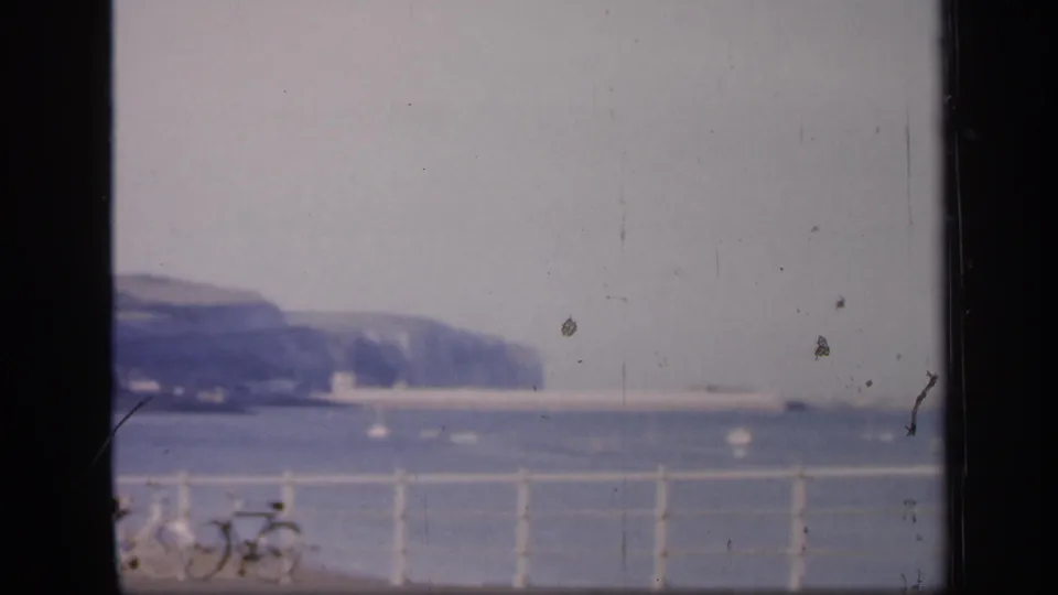 The panning 8mm frame looks past promenade railings and partial bicycles toward the sea and distant chalk coastline.