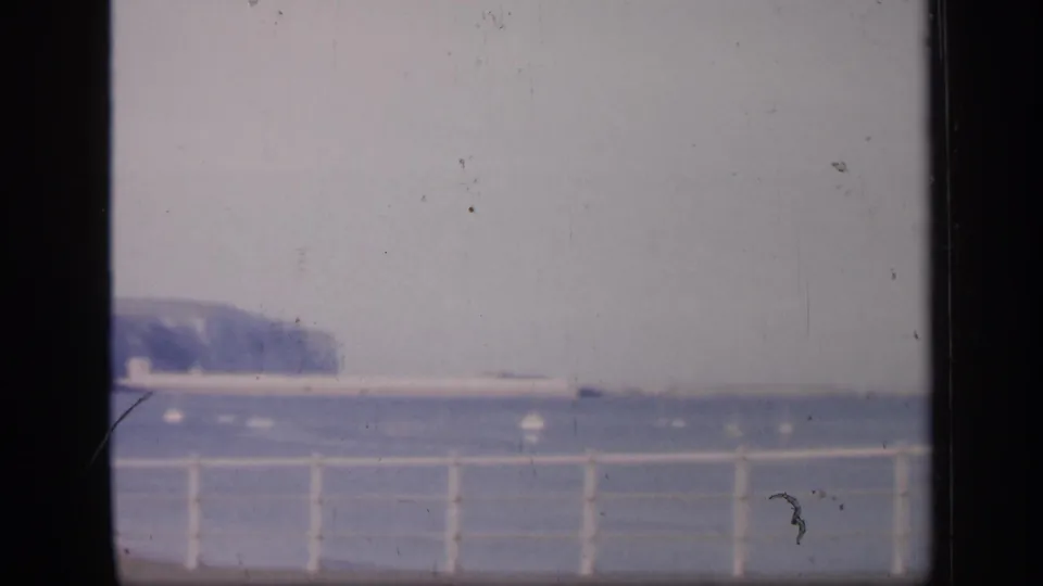 A soft 1949 English seaside frame shows a white railing along the promenade, calm water, and chalk cliffs under haze.