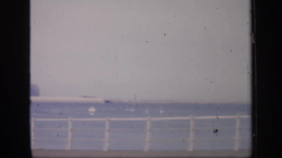 1949 England 8mm frame showing a white waterfront railing, calm sea, pale sky, and faint distant horizon through worn film edges.