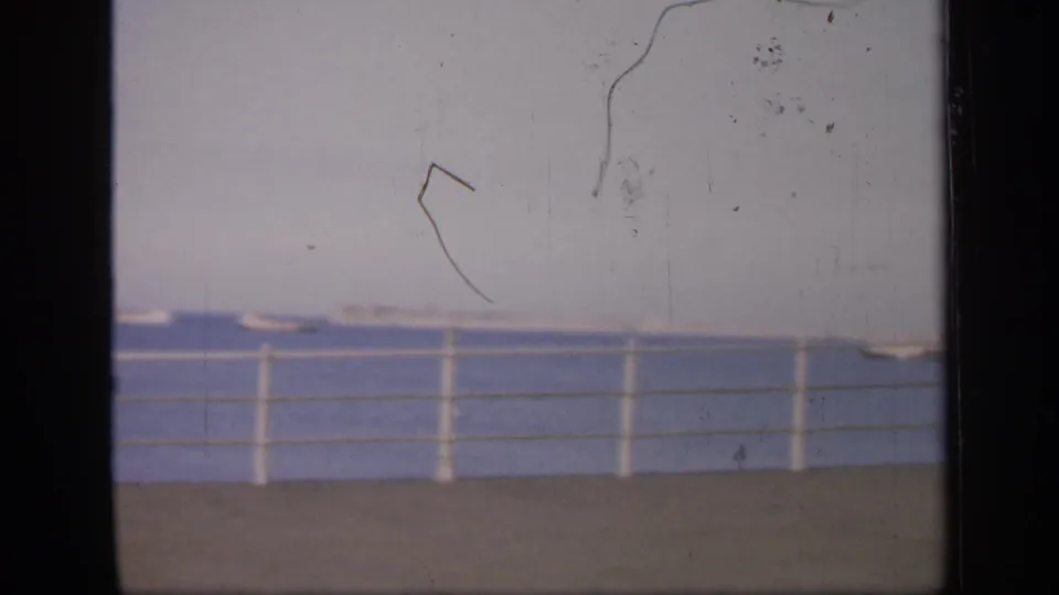 1949 England coastal frame showing open water, a pier railing, distant small boats, pale sky, and prominent surface scratches.