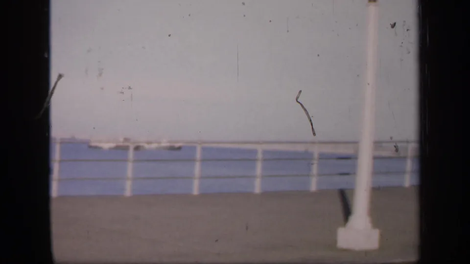1949 England home-movie frame showing water, boats, railings and a white post, with grain and scratches visible.