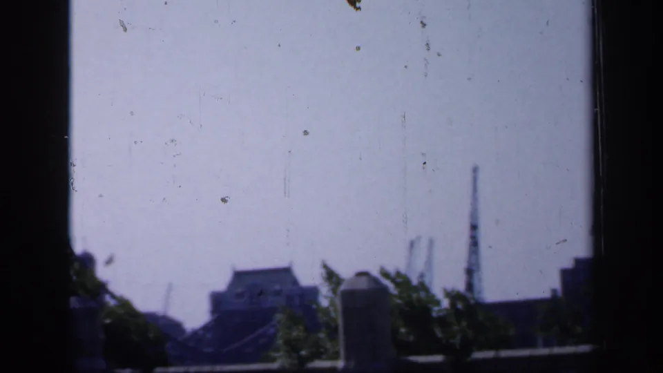 1949 England 8mm frame showing a hazy distant view of Tower Bridge through a dark window frame, with trees and masonry below.