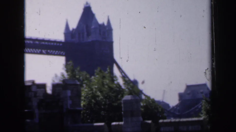 1949 home-movie frame of Tower Bridge with its tower and span visible above trees, masonry posts, and a hazy London skyline.
