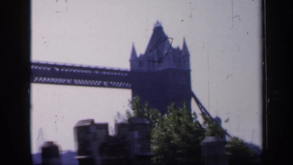 Vintage 8mm frame showing Tower Bridge's span and tower through a dark window edge, with trees and stonework in the foreground.
