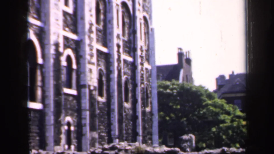 1949 England frame looking past a dark vertical edge toward arched stone windows, trees, and neighboring rooftops.