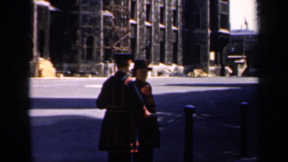 1949 England home-movie frame showing two red-and-black uniformed guards standing in a sunlit courtyard outside a historic stone building.