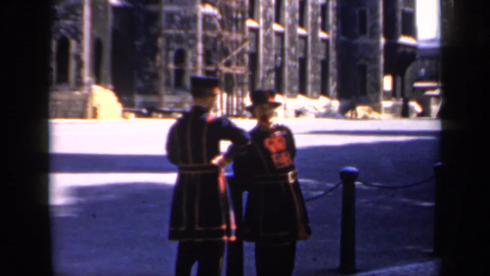 1949 England frame showing uniformed guards beside chained bollards, with a large historic stone building across the sunlit courtyard.