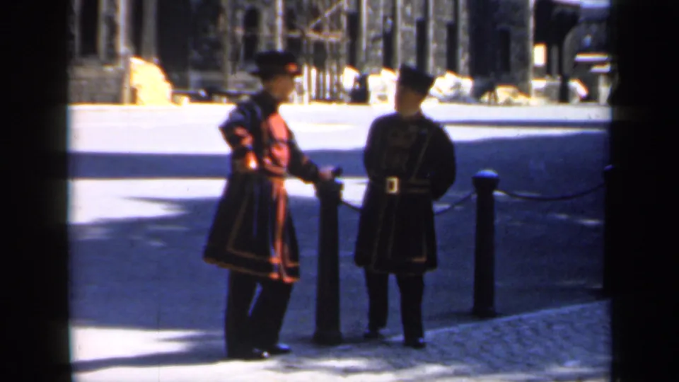 Two Yeomen Warders in ceremonial dress face one another near chained bollards outside the likely Tower of London.