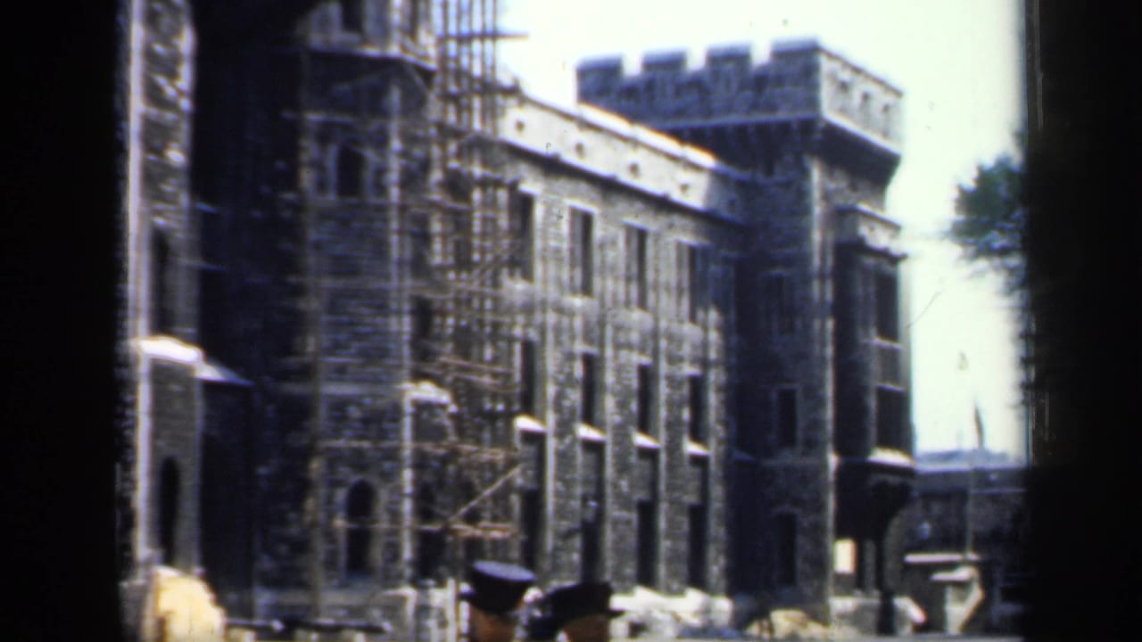 Stone building with crenellated tower, scaffolding, and two hat-wearing figures at the lower edge.