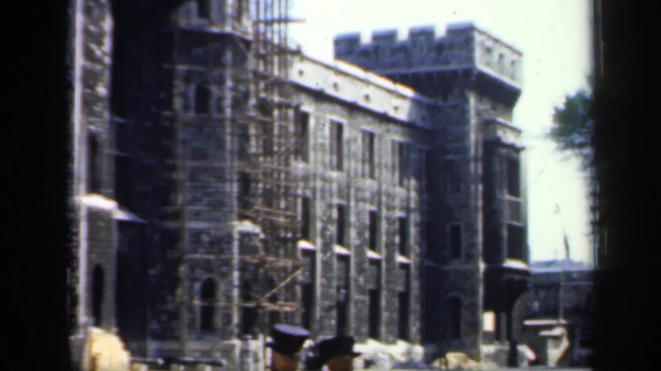 1949 England 8mm frame showing a historic stone building with scaffolding, arched windows, and two foreground figures in dark hats.