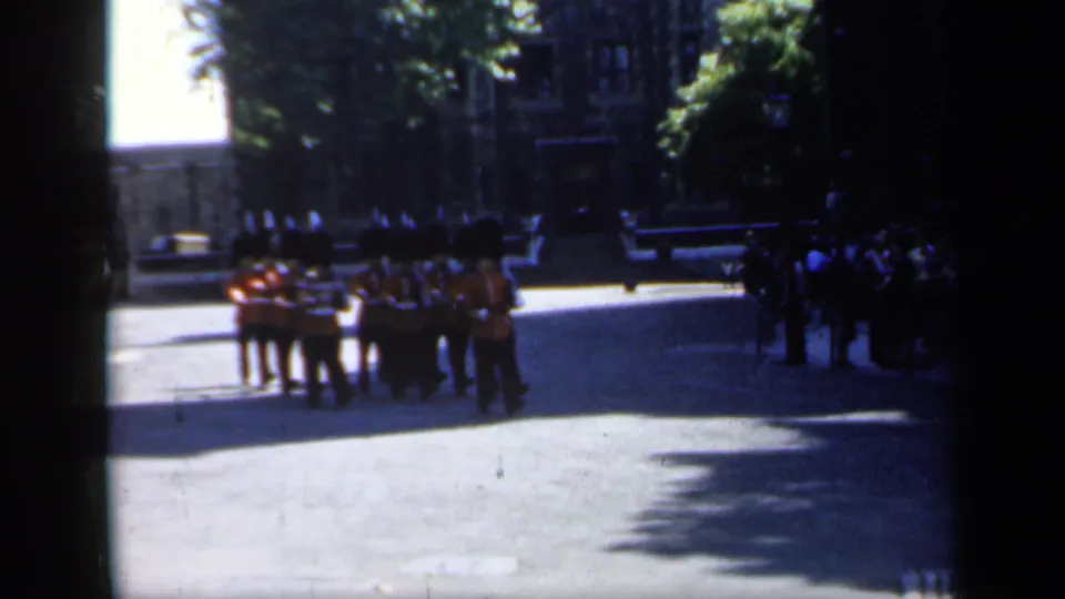 British soldiers in red coats and dark bearskin-style hats march in formation through a sunlit historic setting in England.