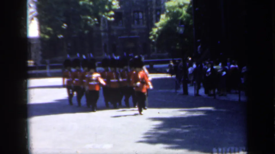 Ceremonial soldiers in red coats and dark hats march past a line of spectators in England, captured with soft 8mm home-movie texture.