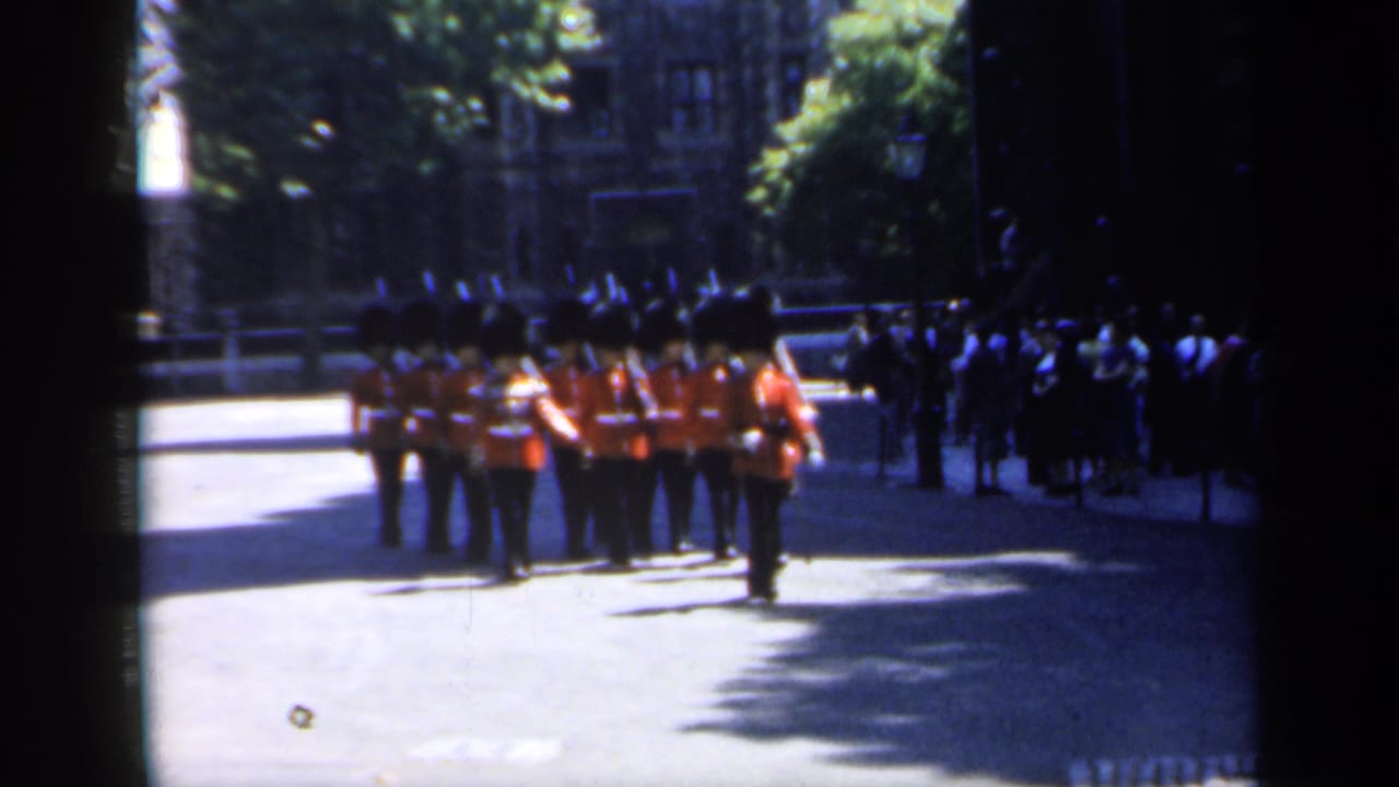 Guards in red tunics march in formation past spectators on a shaded street.