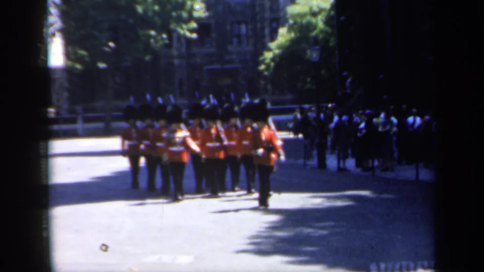 1949 England home-movie frame showing guards in red tunics and tall black hats marching past onlookers on a sunlit street.