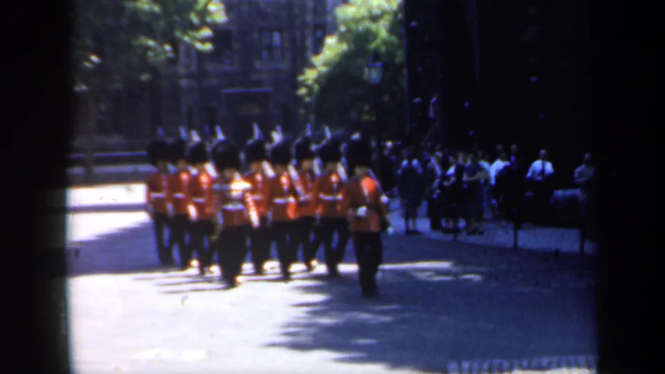 1949 England frame with red-coated guards marching across a bright street, with shaded spectators and stone buildings behind them.