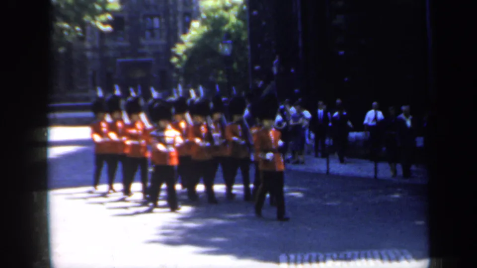 1949 England home-movie still showing ceremonial guards in red tunics passing spectators under strong sunlight and shadow.