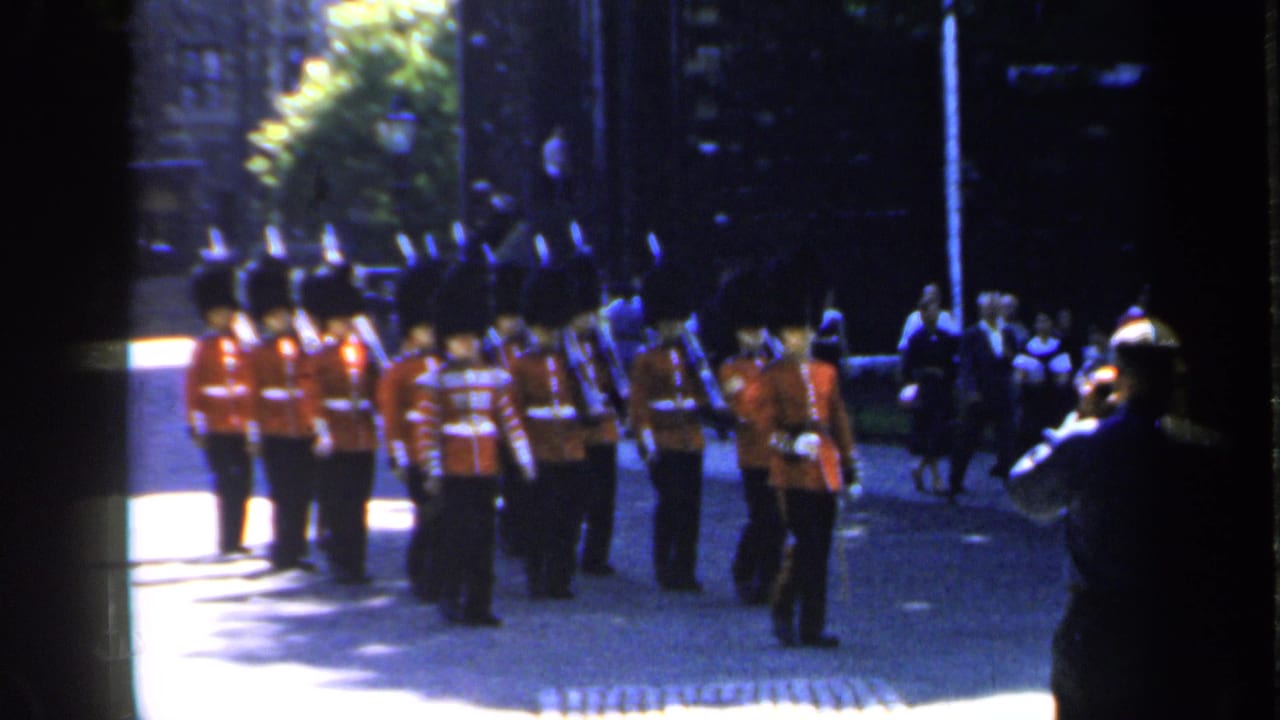 British guards in red tunics stand and march on a cobblestone road as spectators watch nearby.