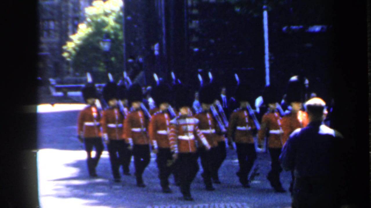 Rows of red-uniformed guards march past a standing spectator on a cobblestone street.