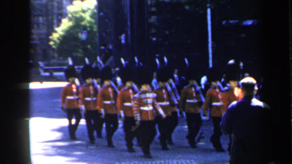 1949 England frame of British ceremonial guards marching in formation past a spectator, with dark buildings and sunlit paving behind them.