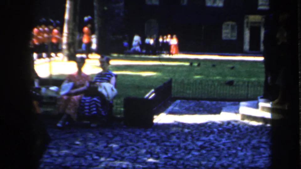 1949 England home-movie view of seated visitors near a lawn and cobblestones, with red-uniformed guards visible in the distance.
