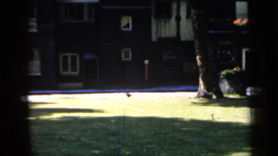 1949 England 8mm frame showing a quiet courtyard with dark building fronts, windows, a bright lawn, and a large tree.