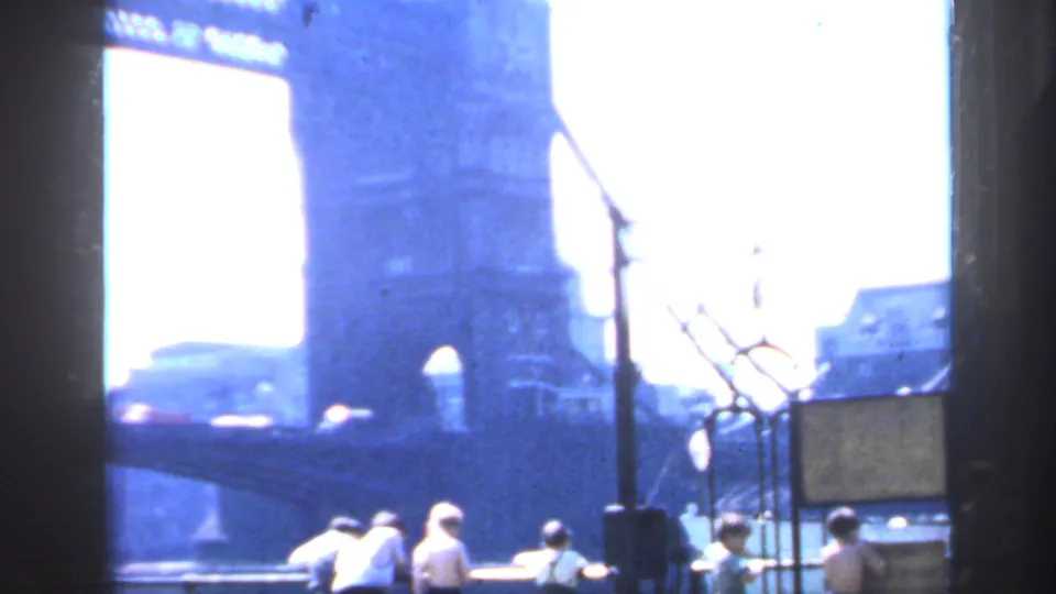 1949 London home-movie frame showing Tower Bridge from a riverside position, with railings, a signboard, and small distant figures.