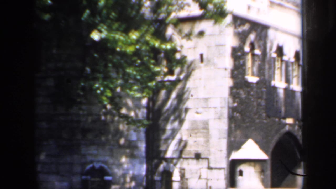 1949 England home-movie view of a stone building facade with windows, trees, and dark foreground obstruction.