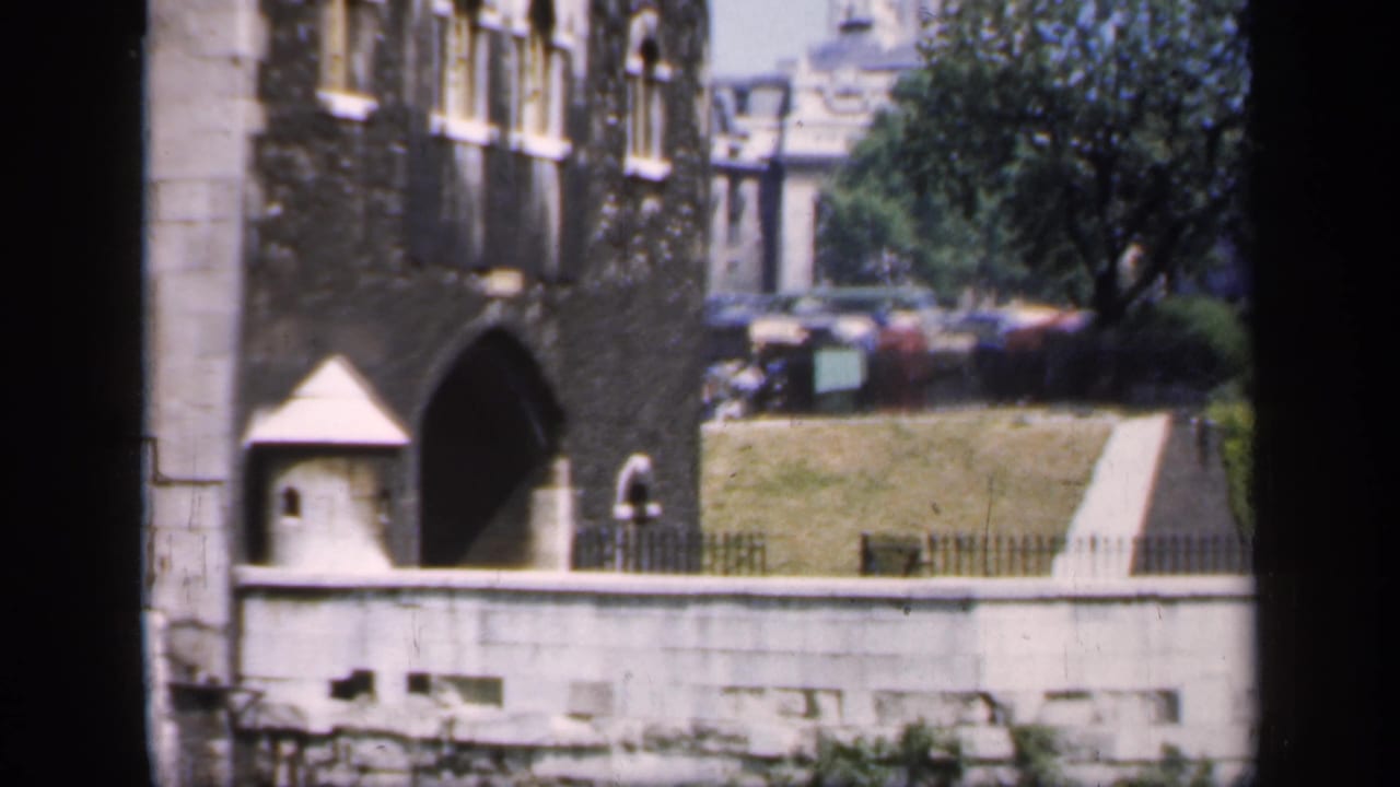 1949 England exterior with a historic arched entrance, low stone wall, lawn, trees, and distant parked cars.